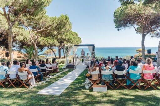 An elegant, luxurious wedding ceremony setup in the manicured Taboo Gardens of Pine Cliffs Resort, featuring white chairs, floral arch, and crisp linens. Luxury outdoor wedding ceremony with white chairs and large floral arch in the manicured Taboo Gardens at Pine Cliffs Resort, Algarve, Portugal.